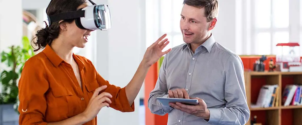 A woman wears a VR headset while a man takes notes of her actions on a tablet.