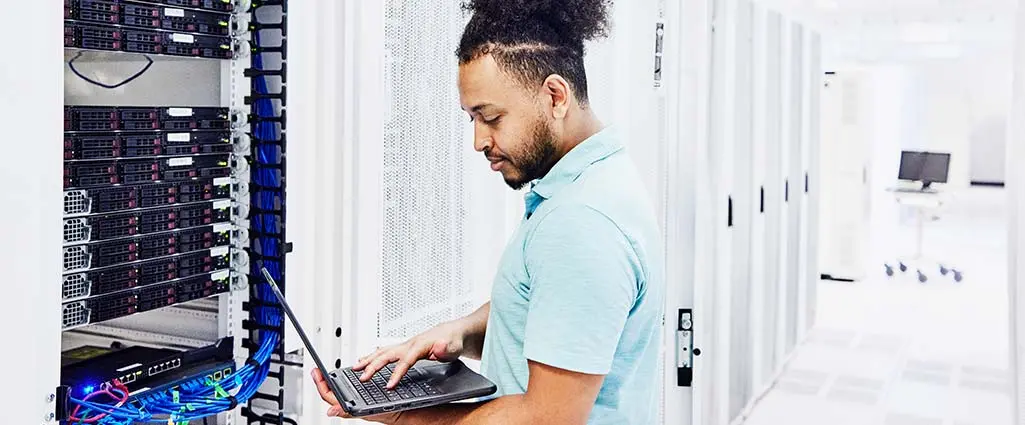 A man in a datacenter working on a server while holding a laptop